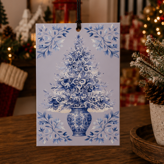 A square Christmas ornament hanging from a black cord, featuring a blue and white illustration of a decorated Christmas tree in a vase, set on a wooden table with a warm, festive background.