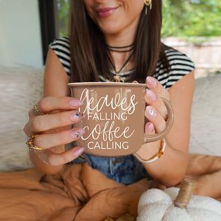 leaves falling coffee calling cup with pumpkin bread
fall countertop decor featuring brown quote mug
autumn coffee bar setup with leaves and coffee mug
brown fall mug styled with beads and greenery
leaves falling coffee calling mug by lit fall candle