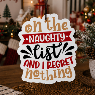 A festive Christmas ornament or wall sign with the text 'on the NAUGHTY list AND I REGRET nothing' in tan, red, and black lettering on a white die-cut background, hanging in a decorated holiday room with stockings, a Christmas tree, and warm string lights.